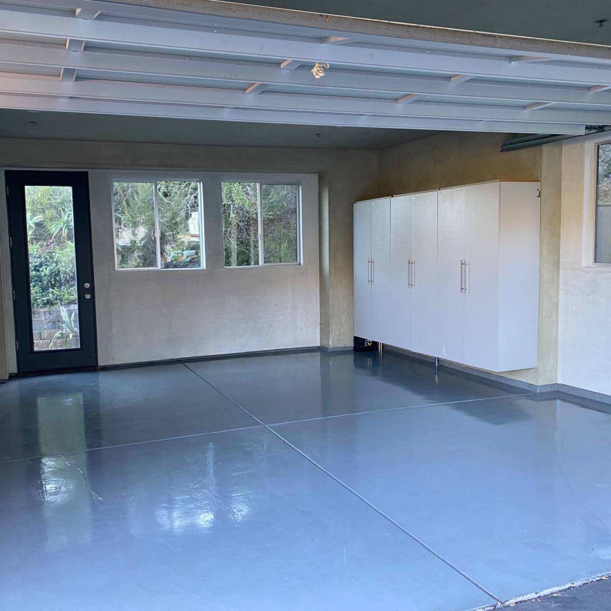 A clean, empty garage with a smooth concrete floor, a large window, a door, and white storage cabinets against the wall.