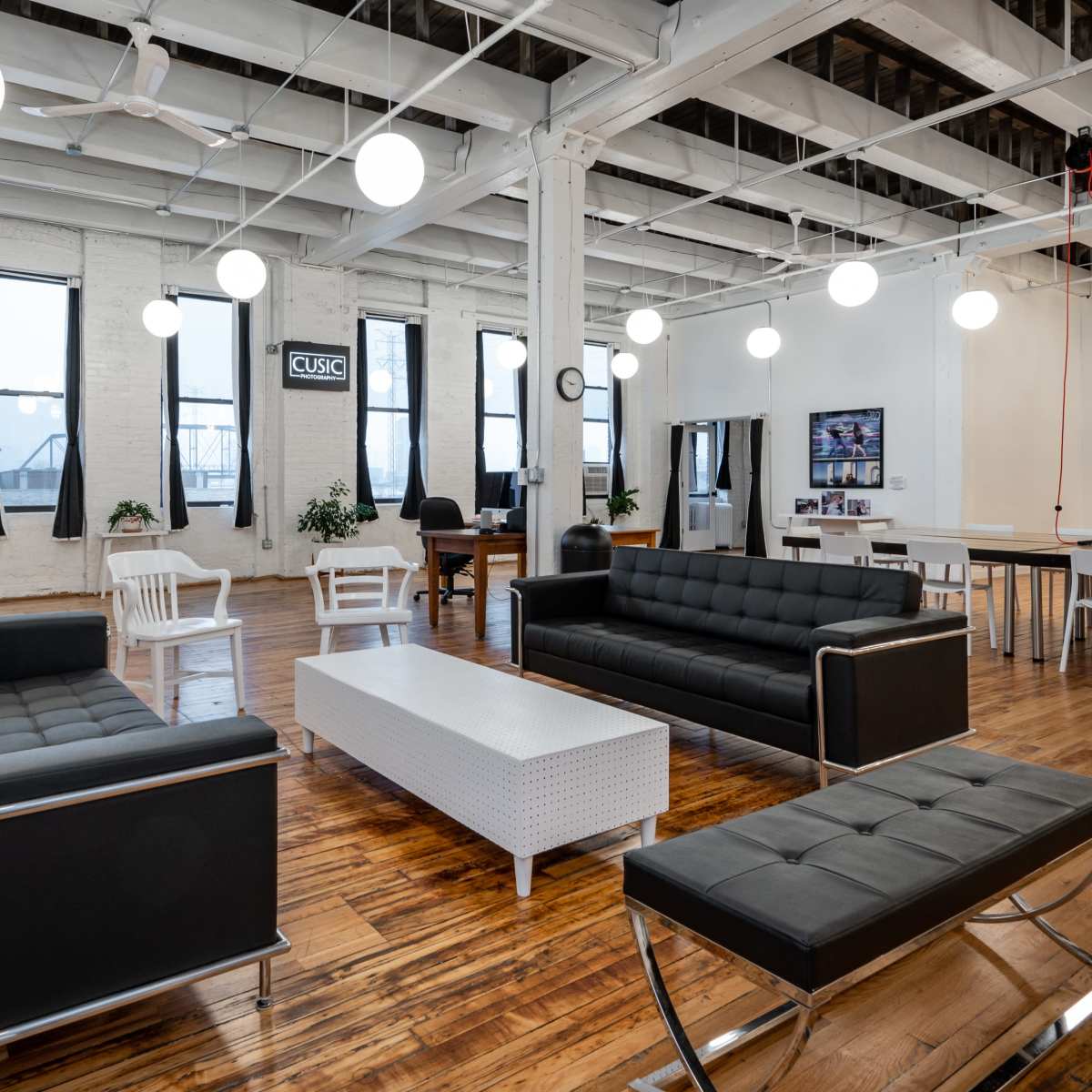 A modern, spacious lounge area with black sofas, a white coffee table, and a dining table surrounded by large windows and hanging lights.