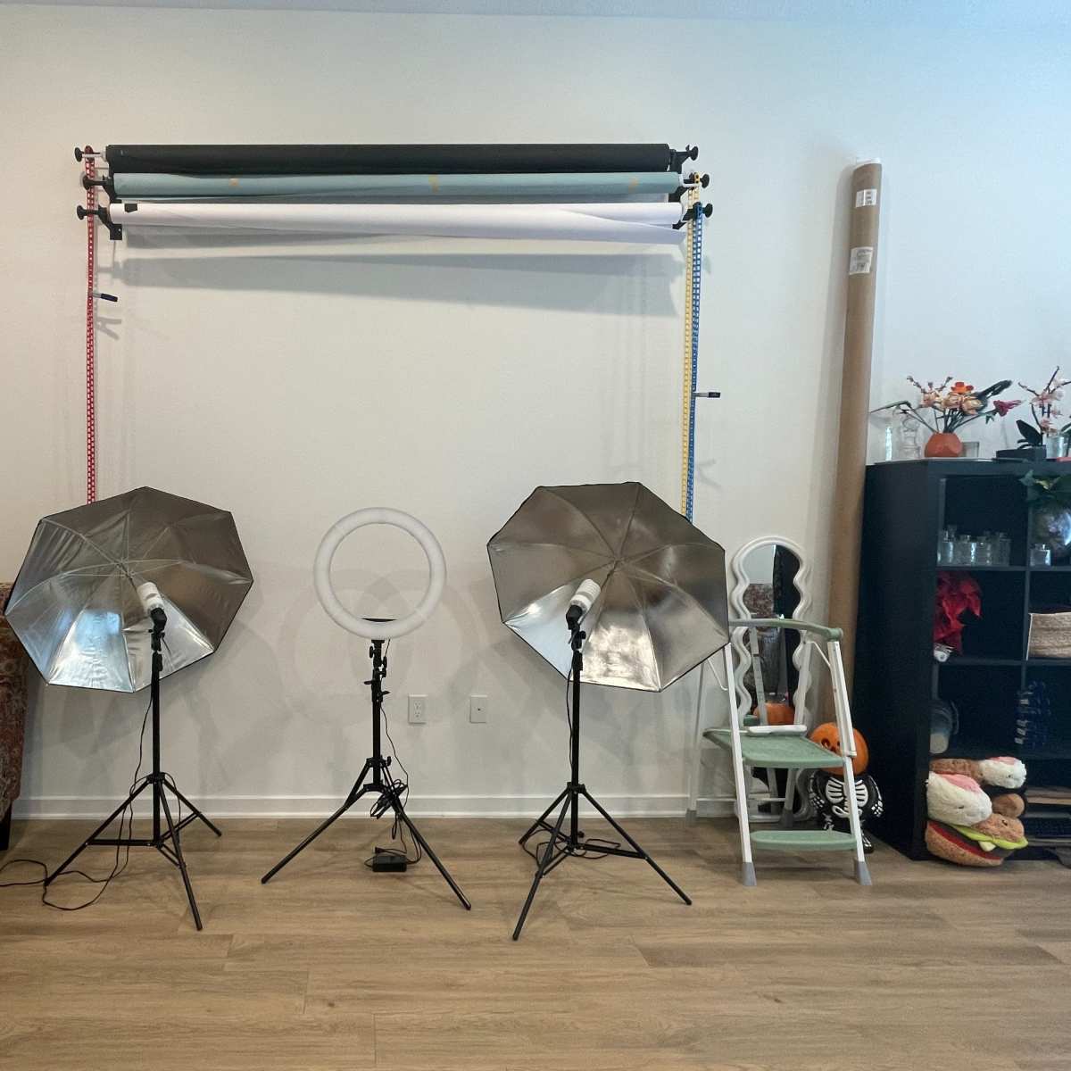 A photography setup featuring two softbox lights and a ring light positioned in front of a backdrop with rolls of paper, alongside a decorative chair and a shelf with various items.