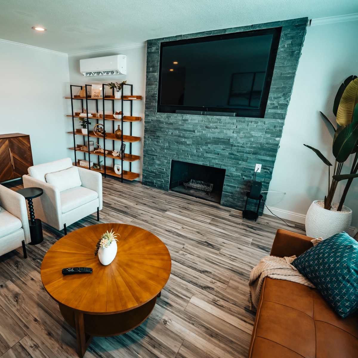 A modern living room with two white upholstered chairs, a brown leather sofa, a round wooden coffee table, and a stone fireplace featuring a mounted television above it.