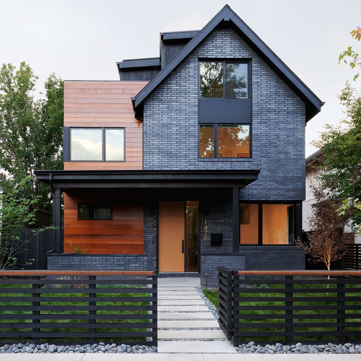 A modern two-story house with a dark brick facade, wooden accents, and a landscaped front yard featuring a black slatted fence and a pathway leading to the entrance.