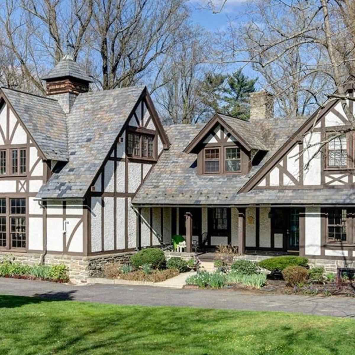 A large, two-story house with a steeply pitched roof, exposed wooden beams, and a stone foundation, surrounded by greenery.