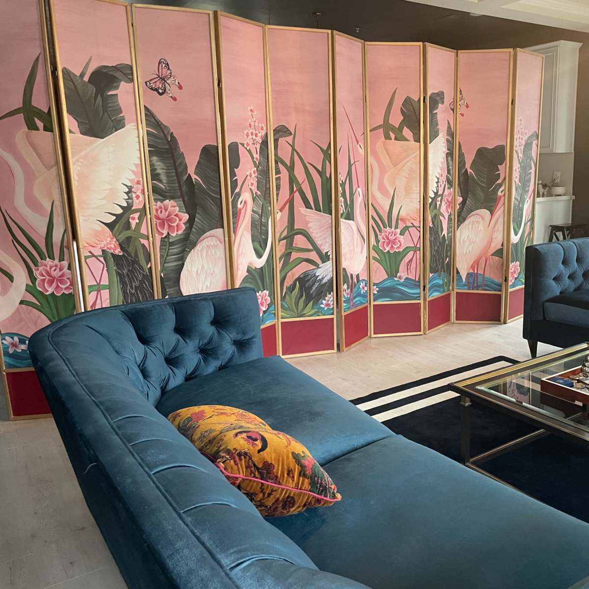 A living room with two blue velvet sofas facing a glass coffee table, set against a decorative screen featuring pink and white cranes surrounded by tropical plants.