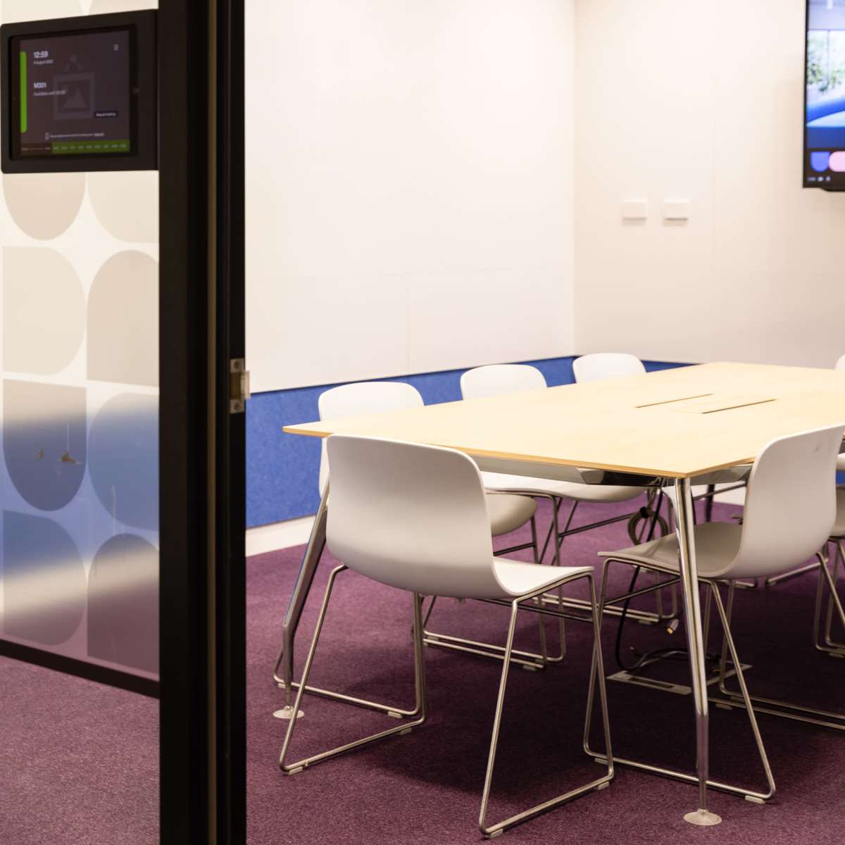 A modern conference room with a large wooden table surrounded by white chairs, featuring a digital display on one wall.