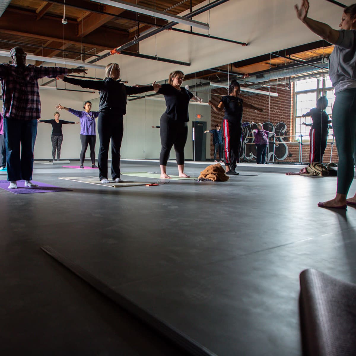 A group of people is practicing yoga in a spacious studio with large windows and wooden beams overhead.