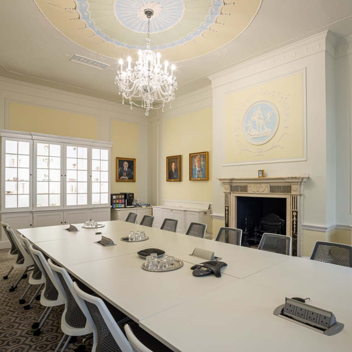 A formal conference room with a long white table, black and white chairs, a chandelier, and framed portraits on the walls.