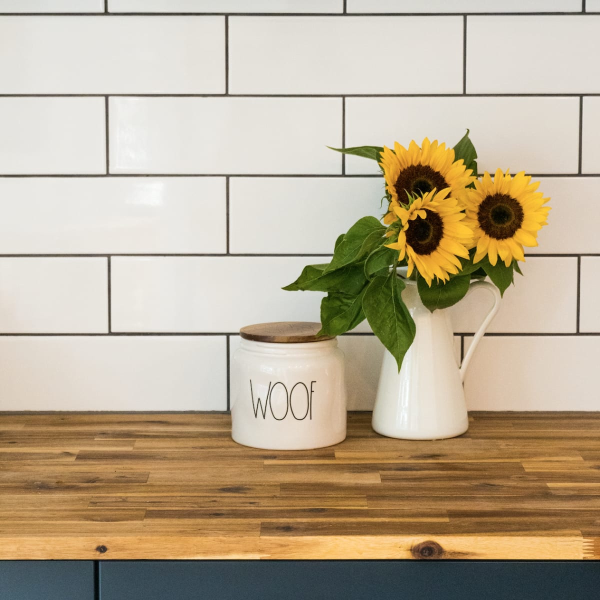 A kitchen counter with a wooden surface featuring a white canister labeled "WOOF" and a white pitcher holding sunflowers, against a backdrop of navy cabinets and white tiled backsplash.