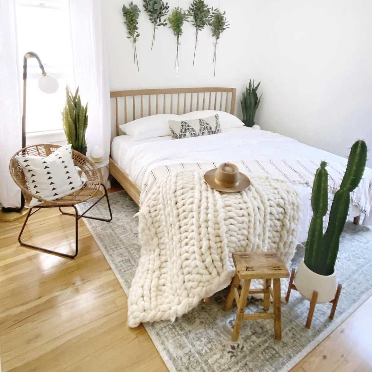 A minimalist bedroom with a bed, a woven chair, and potted plants, featuring a cozy blanket and decorative accents on the walls.