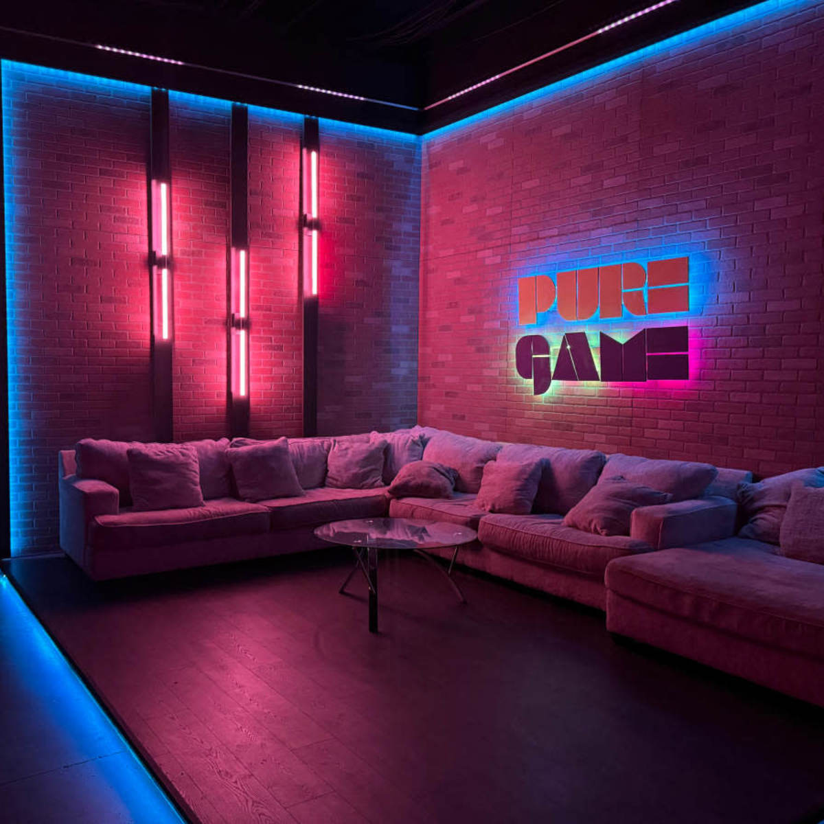 A modern lounge area featuring a large sectional couch, a glass coffee table, and illuminated brick walls displaying the words "PURE GAME" in vibrant colors.