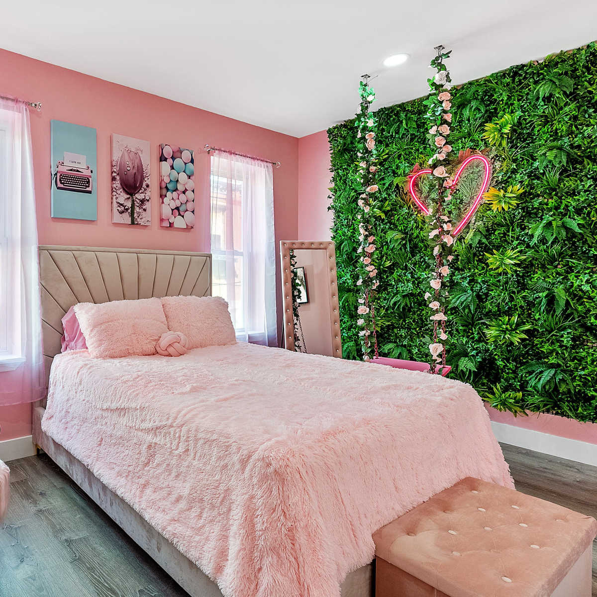 A pastel pink bedroom features a bed with a plush cover, a mirrored vanity table, and a green wall adorned with artificial plants and decorative hearts.