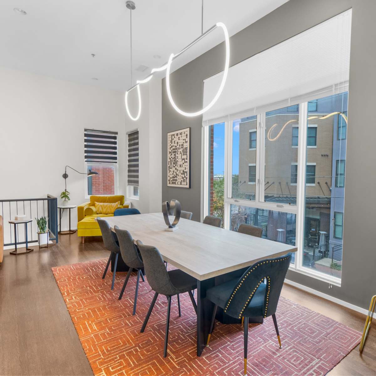 A modern dining area features a sleek table with black chairs, illuminated by contemporary light fixtures, adjacent to a living space with a yellow sofa and large windows.