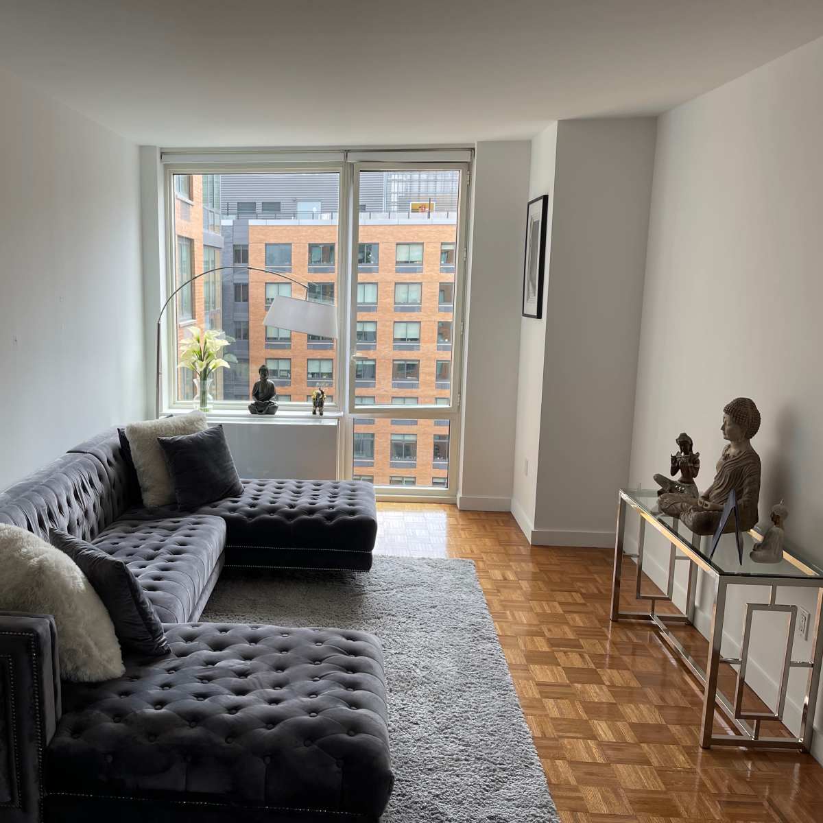 A modern living room features a gray tufted sofa, a decorative rug, and large windows overlooking a cityscape.