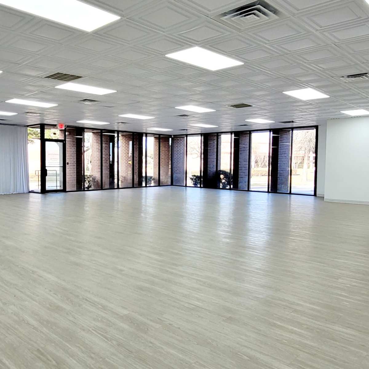 A spacious, empty interior room with a light wood floor, large windows, and white walls.