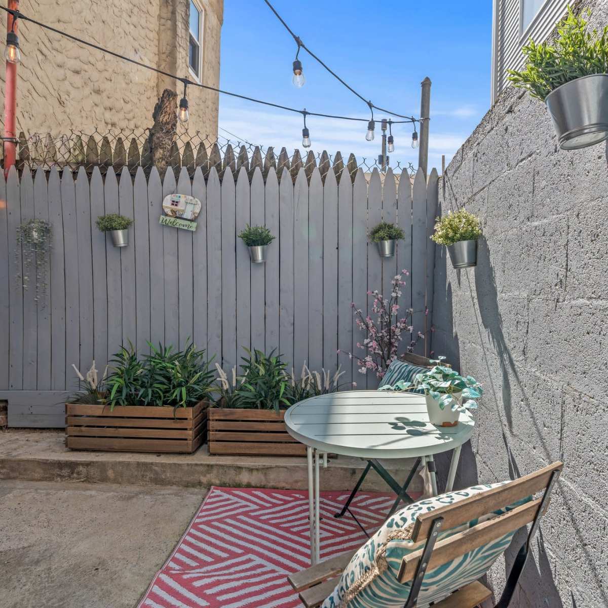 A small outdoor area with a table, a chair, potted plants, and string lights hanging above a gray wood fence.