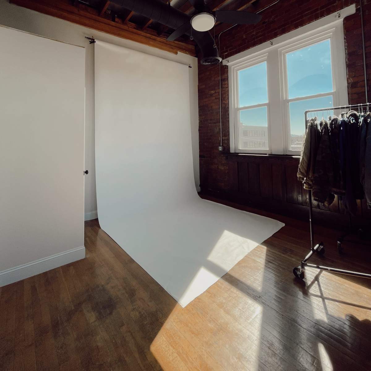 A spacious, well-lit room with a white backdrop, large windows, and a clothing rack against a brick wall.