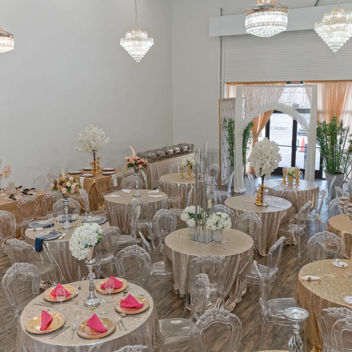 An elegantly arranged banquet hall with round tables covered in gold tablecloths, adorned with floral centerpieces and crystal chandeliers hanging from the ceiling.