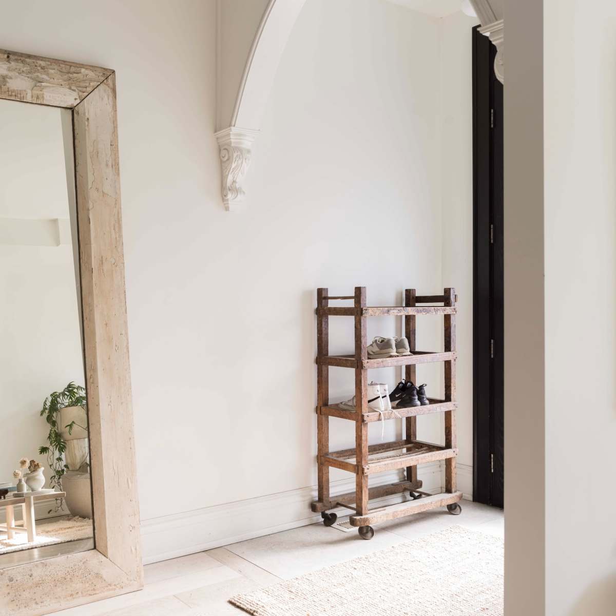 A large, weathered mirror leans against a white wall beside a wooden shoe rack with several pairs of shoes.