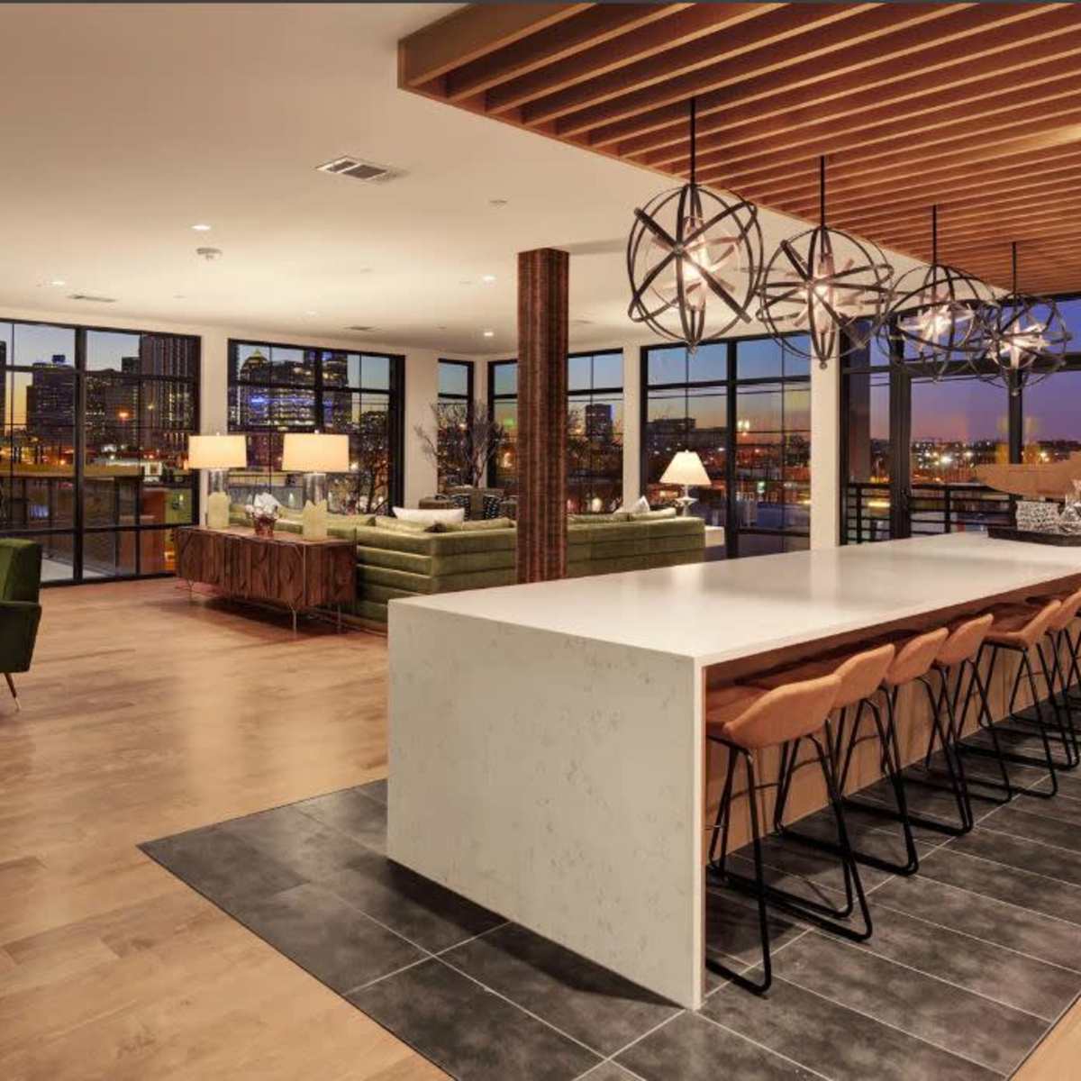 A modern interior space featuring a large white kitchen island with bar seating, green chairs, and large windows revealing a city view at dusk.