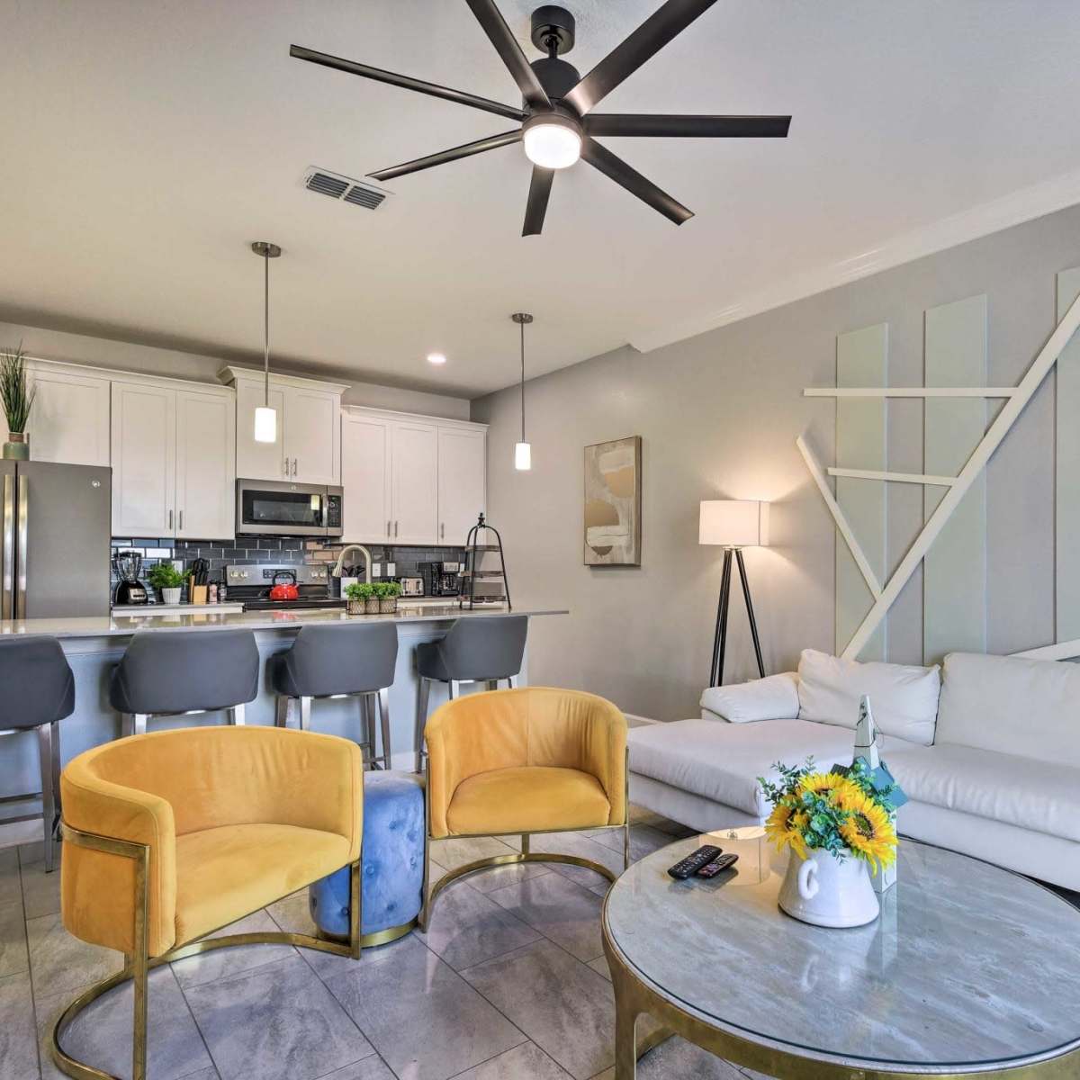 A modern living room features a white sectional sofa, yellow accent chairs, a round coffee table, and an open kitchen in the background.