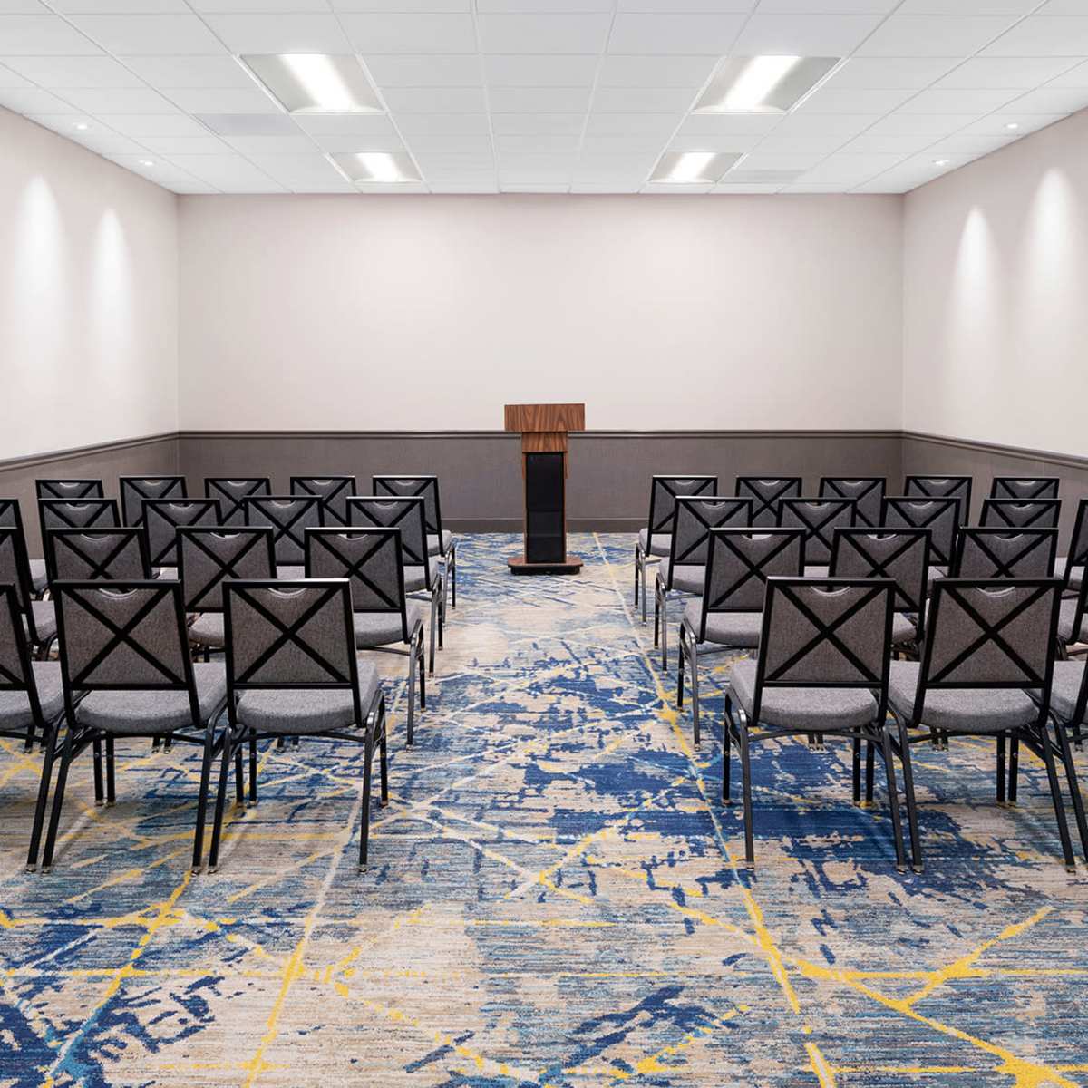 A meeting room features rows of black chairs facing a wooden podium on a patterned carpet.