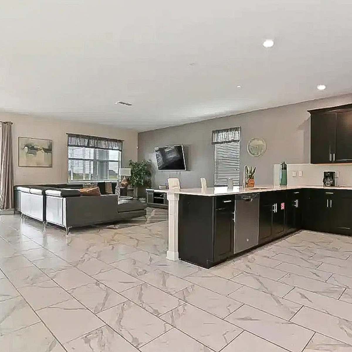A modern open-concept kitchen and living area with tiled flooring, dark cabinetry, and large windows providing natural light.