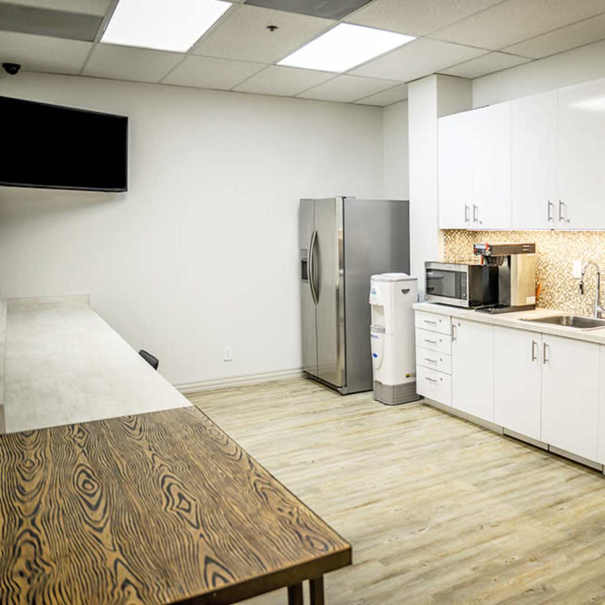 A modern kitchen area with a refrigerator, microwave, sink, and countertop, along with a dining table and wall-mounted television.