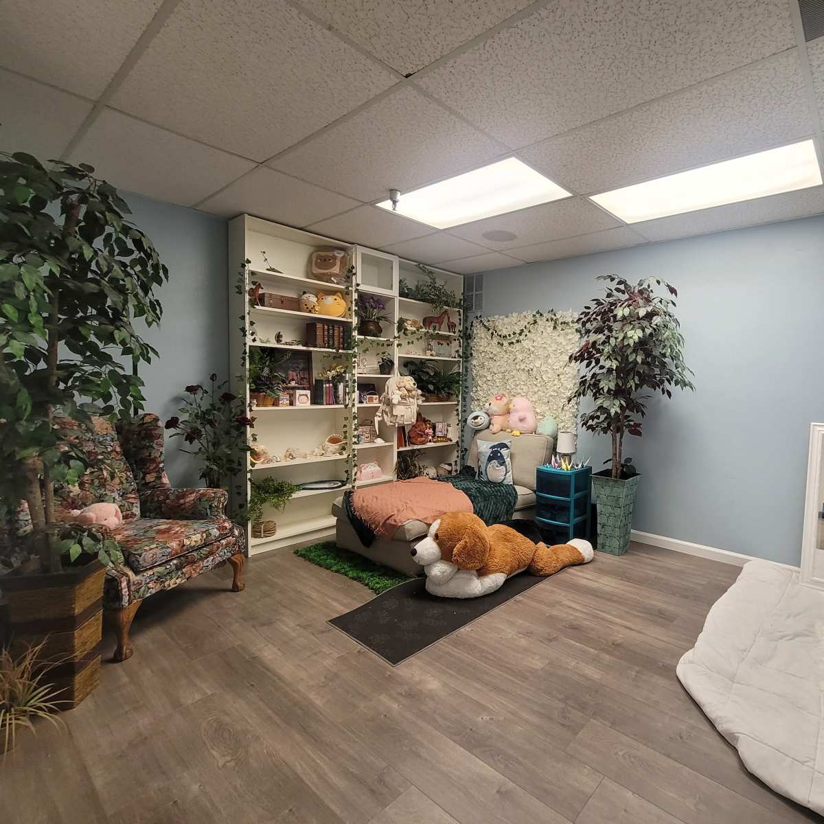 A cozy room with a colorful mural, a shelf filled with various items, a plush chair, a large stuffed animal, and a mirror on the floor near a white blanket.