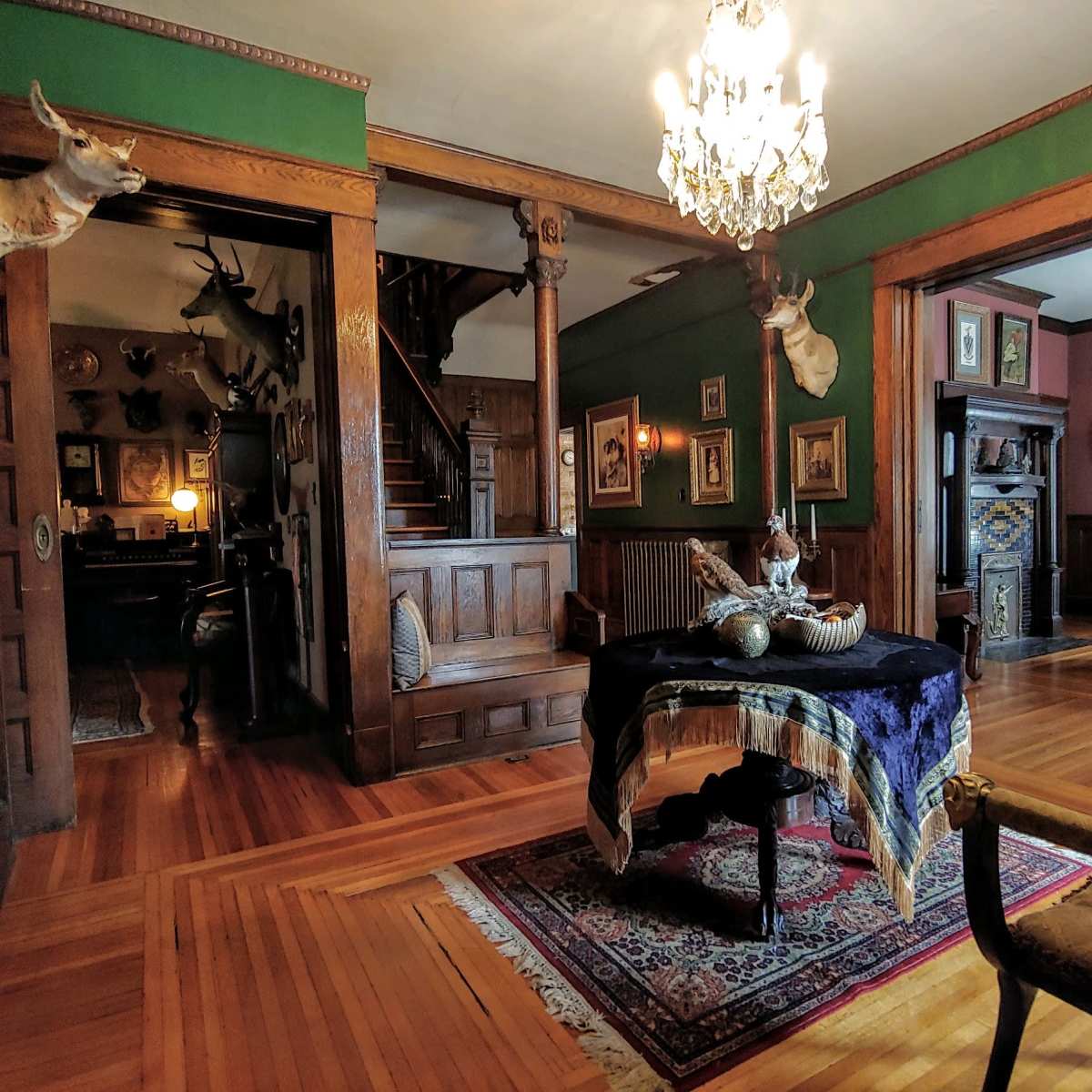 A vintage interior featuring wooden walls, a staircase, and decorative elements including mounted animal heads and ornate furniture.