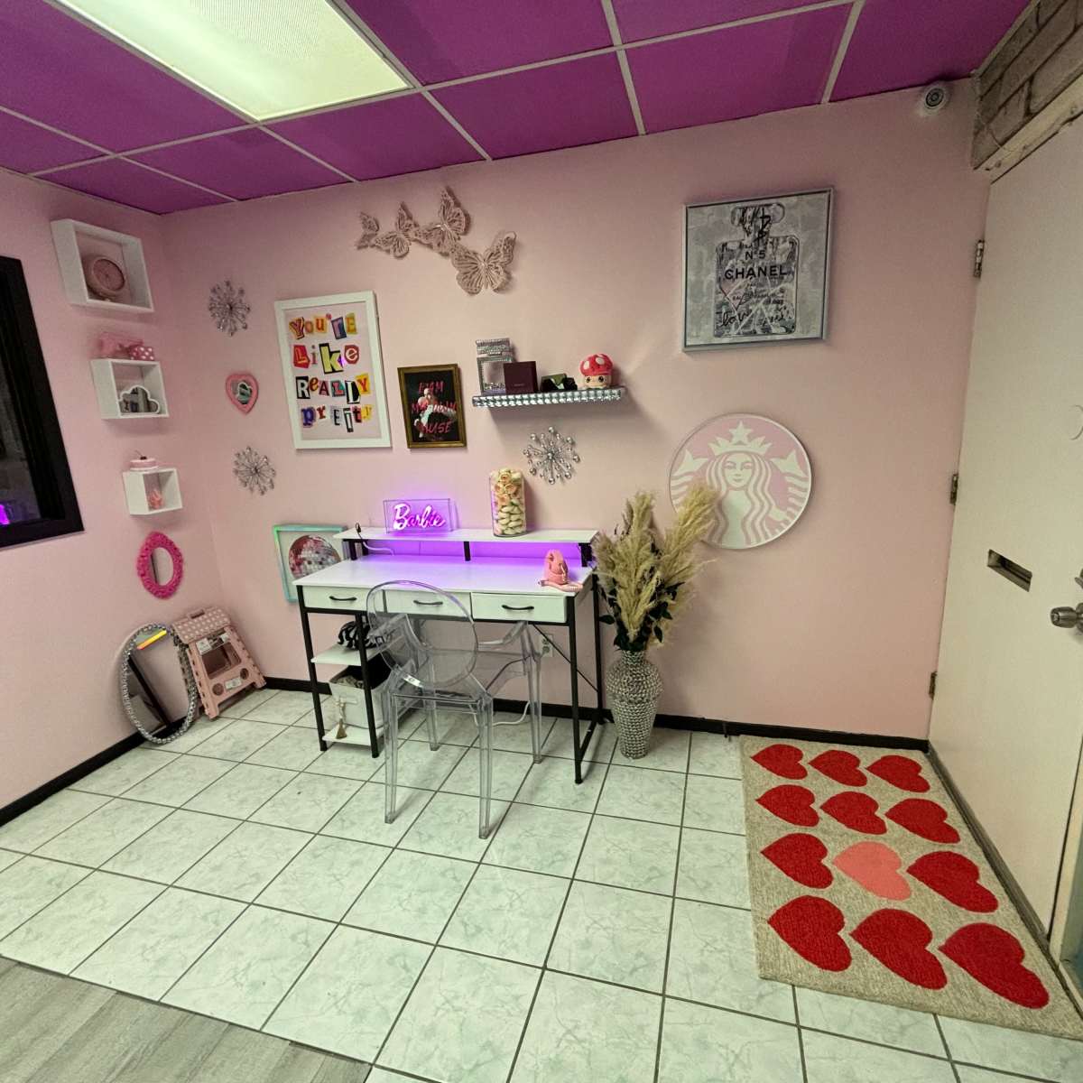 A room with pink walls, a tiled floor, a desk with a light, and decorated with various wall art and a heart-patterned rug.
