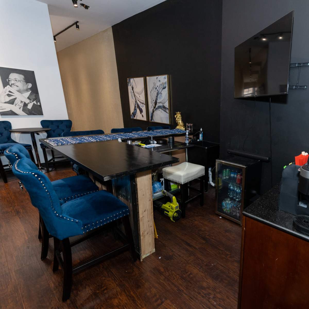 A modern interior space featuring a table with blue upholstered chairs, a wall-mounted television, and a bar area with a drink dispenser and various bottles.