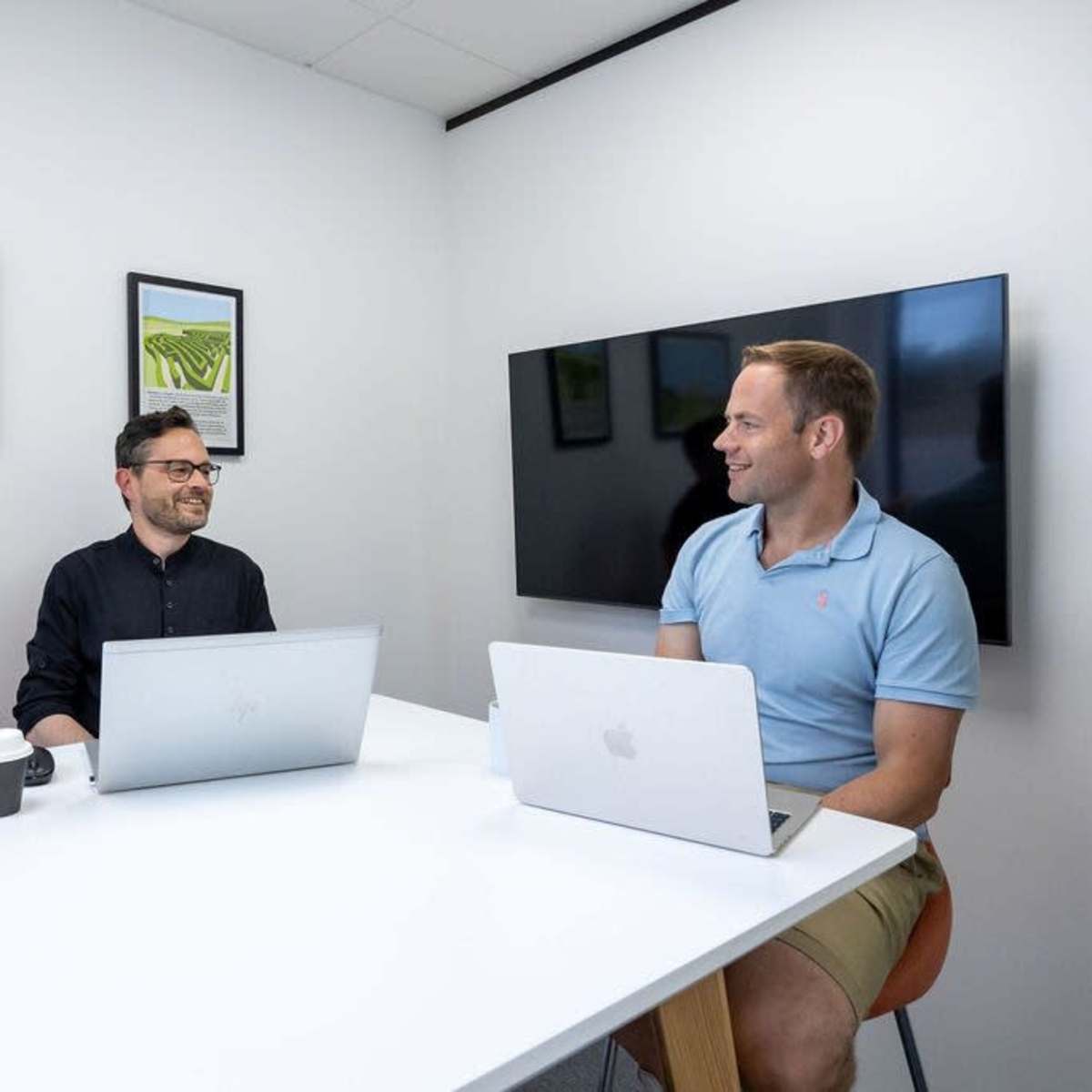 Two men are sitting at a white table in a bright office space, each working on a laptop while engaging in conversation.
