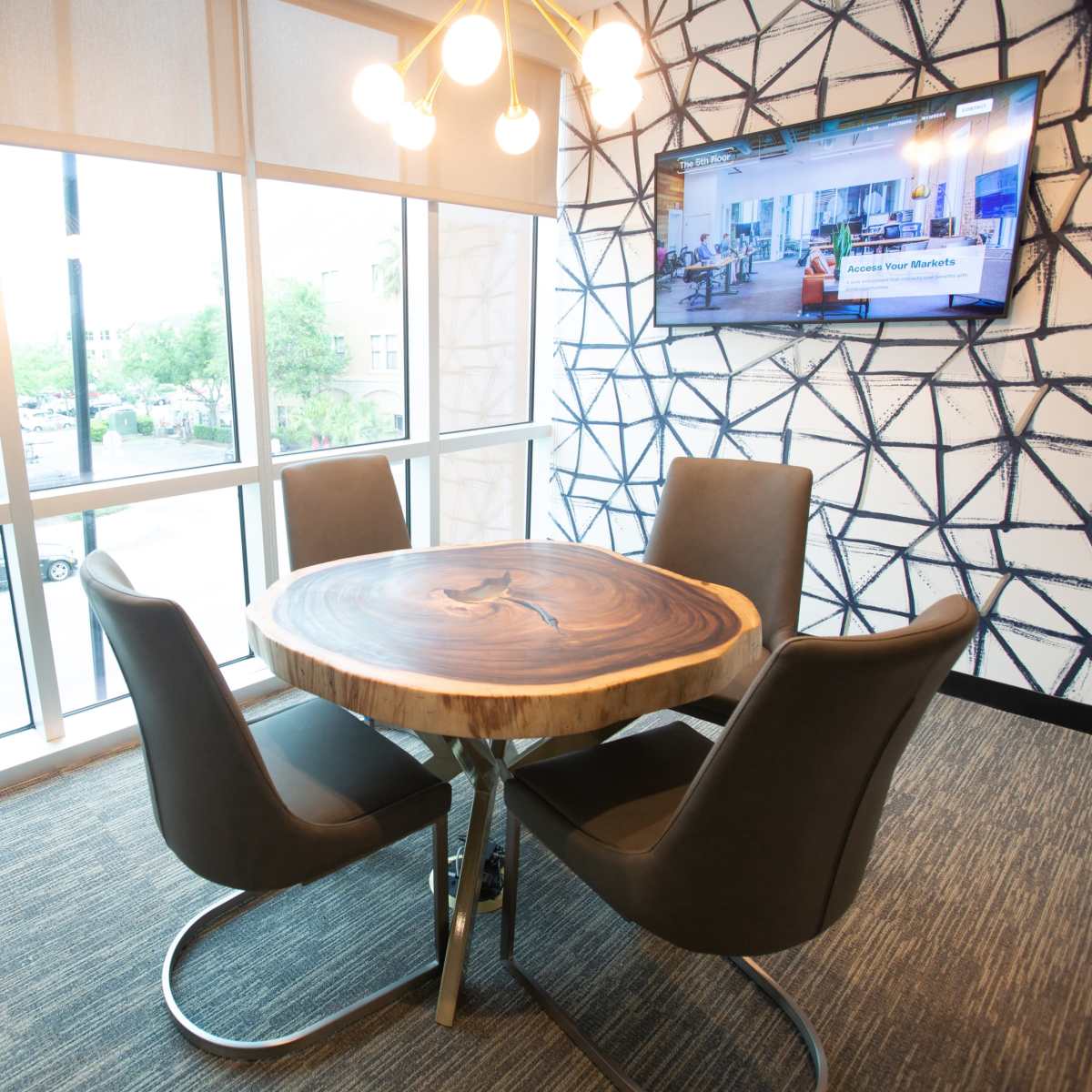A modern meeting area features a round wooden table surrounded by four black chairs, positioned next to large windows displaying an outdoor view.