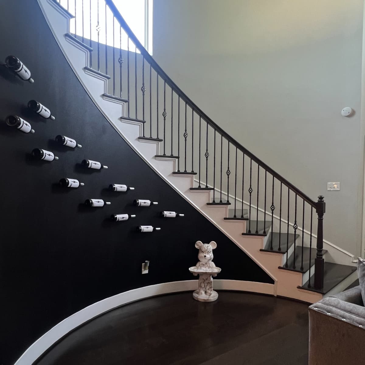 A curved staircase with a black wall displays wine bottles, while a decorative statue stands at the bottom.