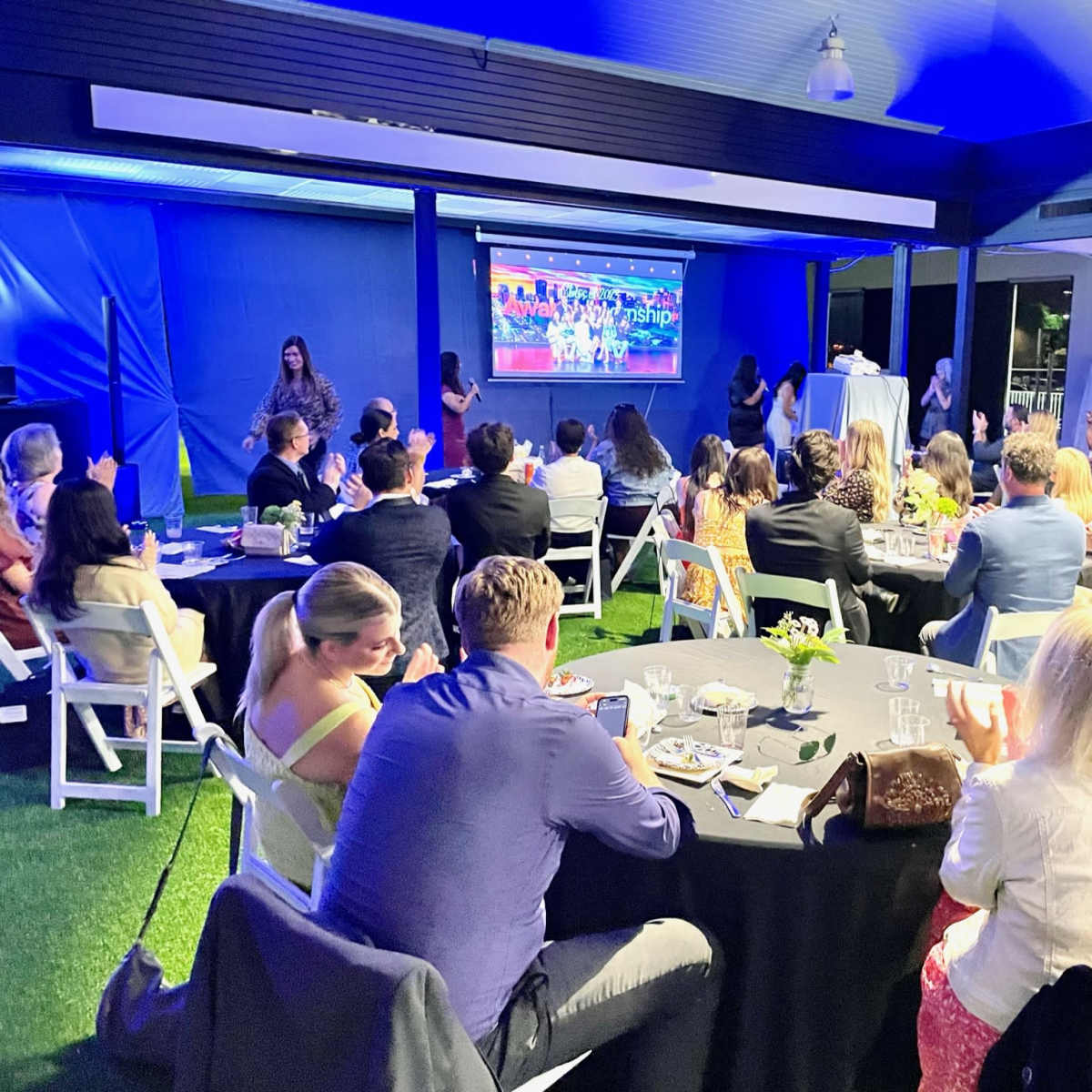 A large group of people is seated at round tables, watching a presentation on a screen against a backdrop of blue lighting.