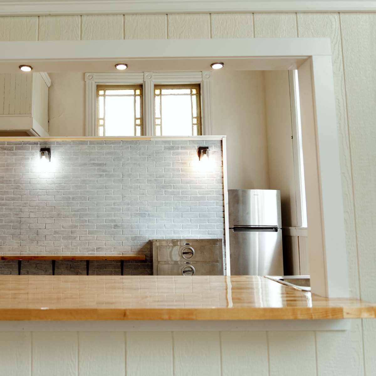 A kitchen with a wooden countertop and a grey brick wall, featuring a stainless steel refrigerator and overhead lighting.