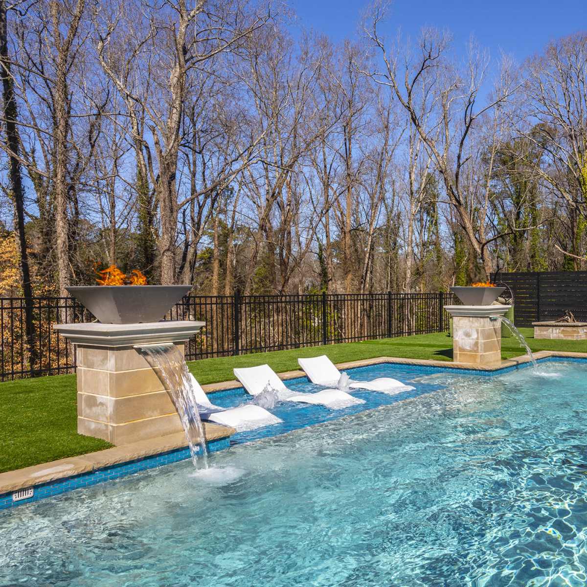 A residential outdoor pool surrounded by landscaped grass, with lounge chairs and a stone wall in the background.