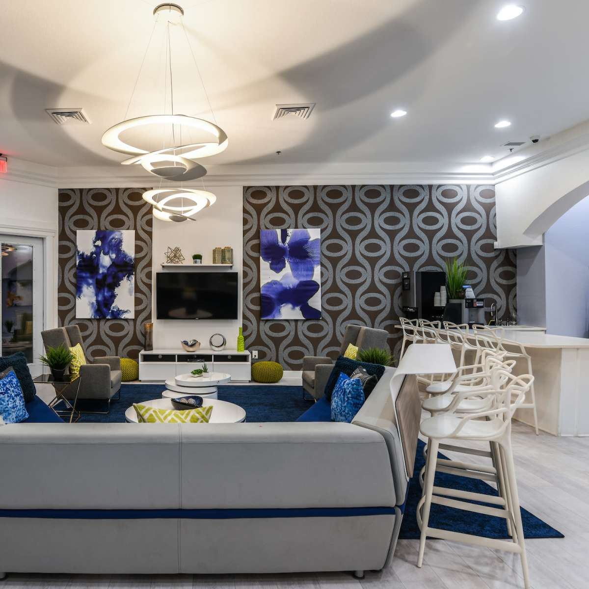 A modern lounge area featuring a gray sectional couch, round coffee tables, and two large artworks on the wall, with a dining space visible in the background.