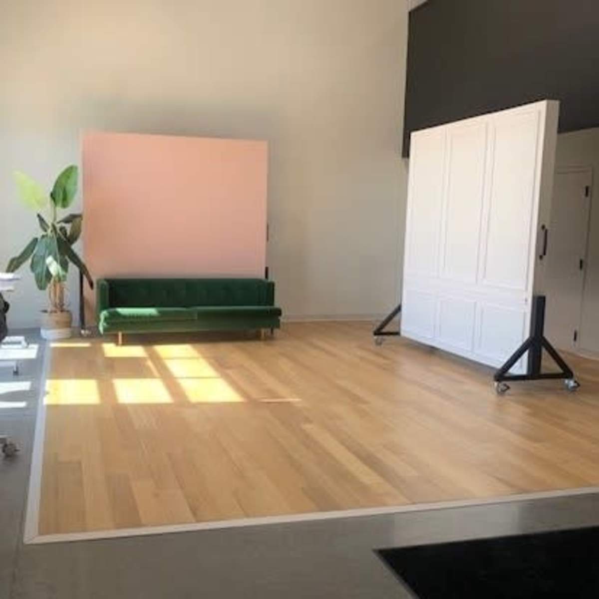A spacious room featuring a green velvet sofa, a pink backdrop, and a large white partition on wheels, with light-colored wooden flooring.