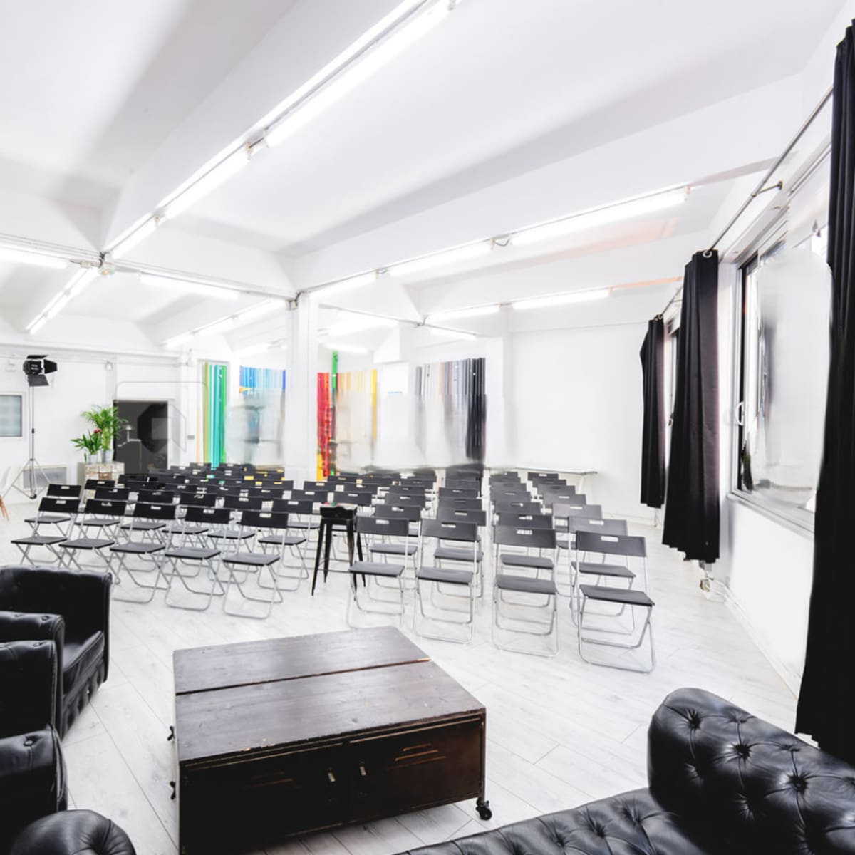 A bright, modern event space features rows of black chairs facing a central area, with a black table and leather sofas in the foreground.