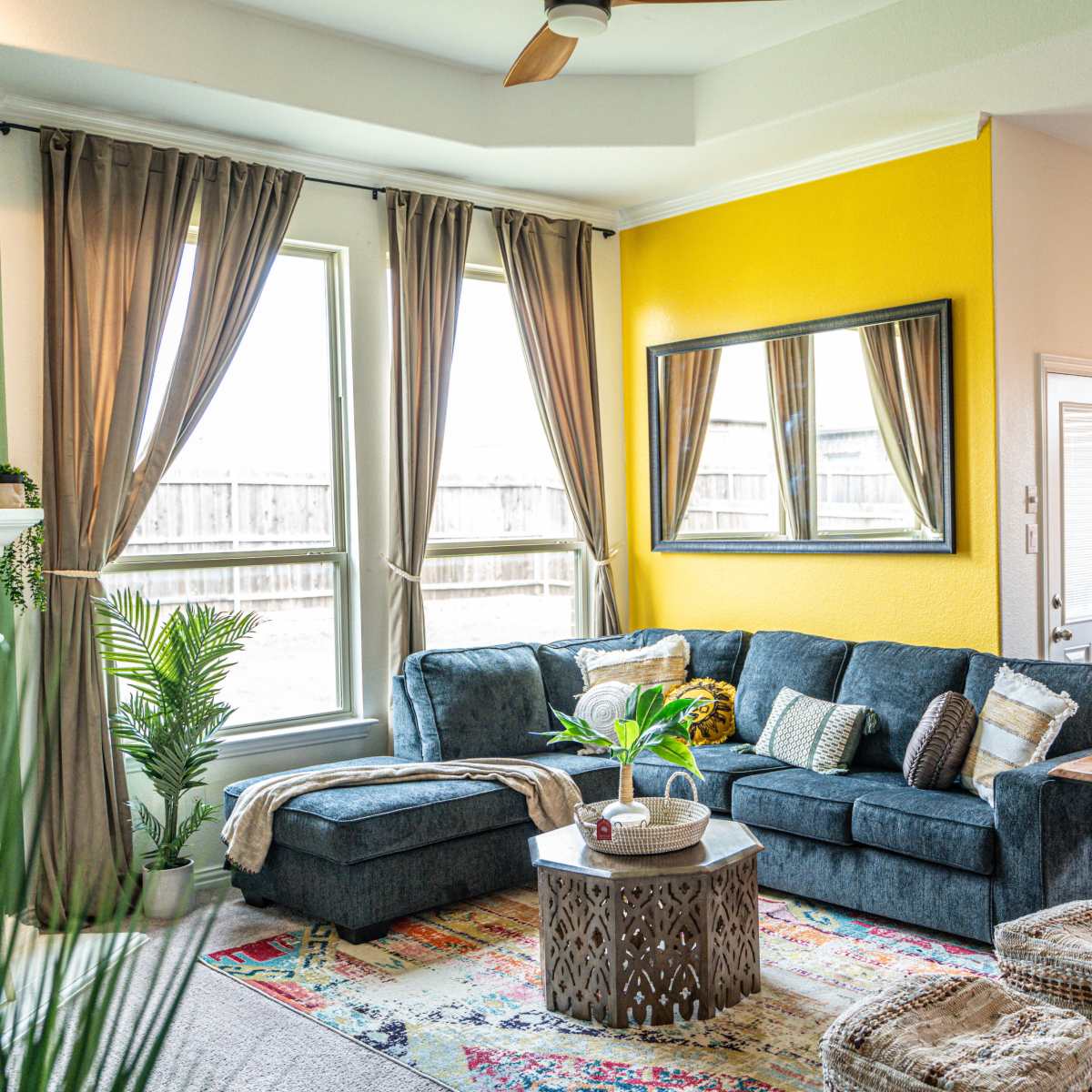 The living room features a blue sectional sofa, a round coffee table, large windows, and a mix of plants and decor against yellow and green walls.