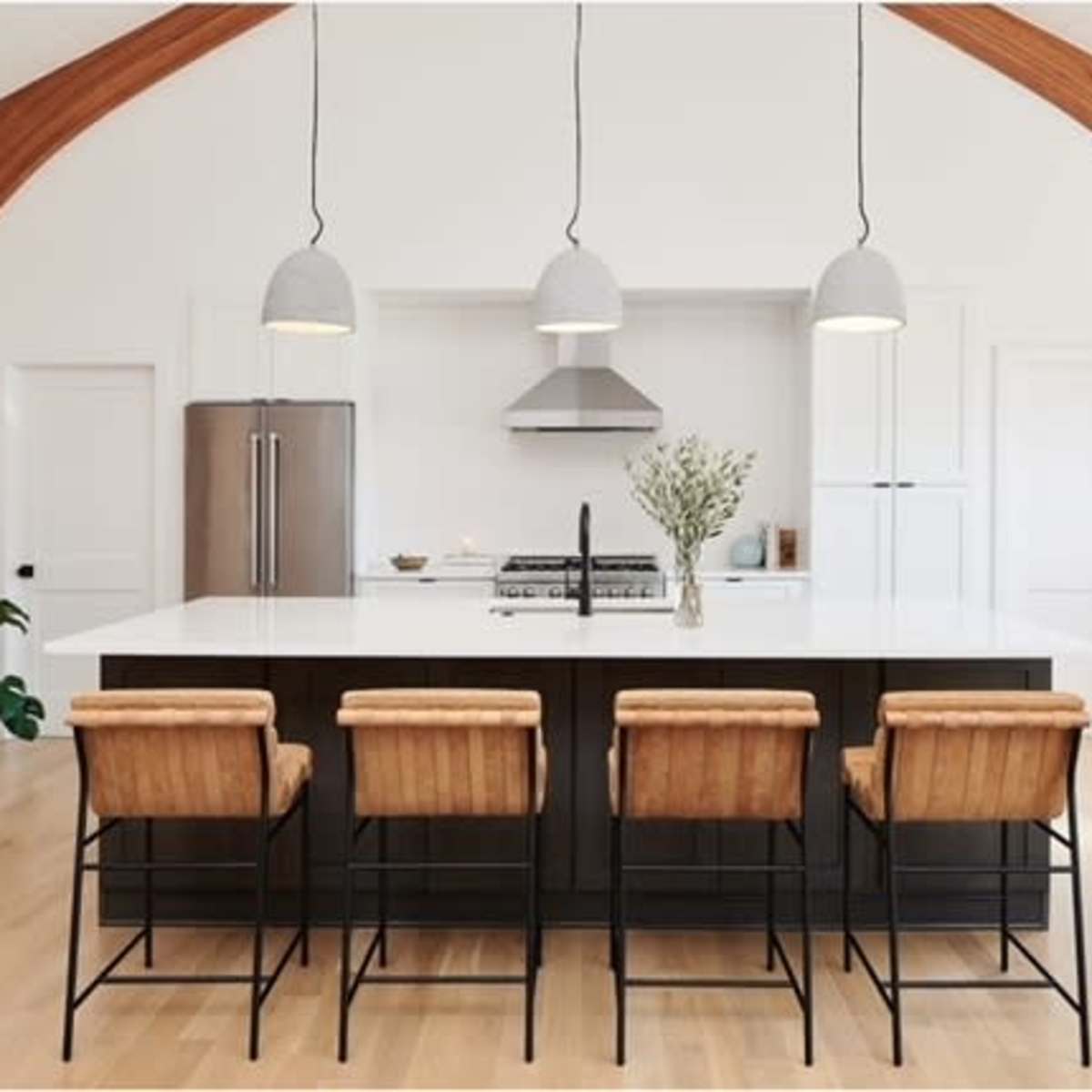 A modern kitchen features a large island with four metal and wood bar stools, pendant lights, and a stainless steel stove beneath a vent hood, set against arched wooden beams.