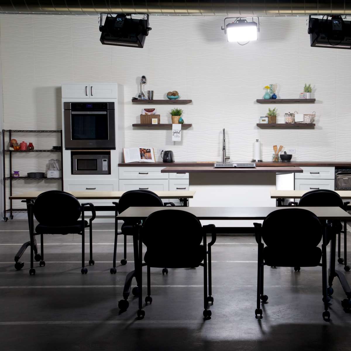 A modern kitchen setup features a stainless steel refrigerator, an oven, and minimalist shelving, with six black rolling chairs positioned around two tables.