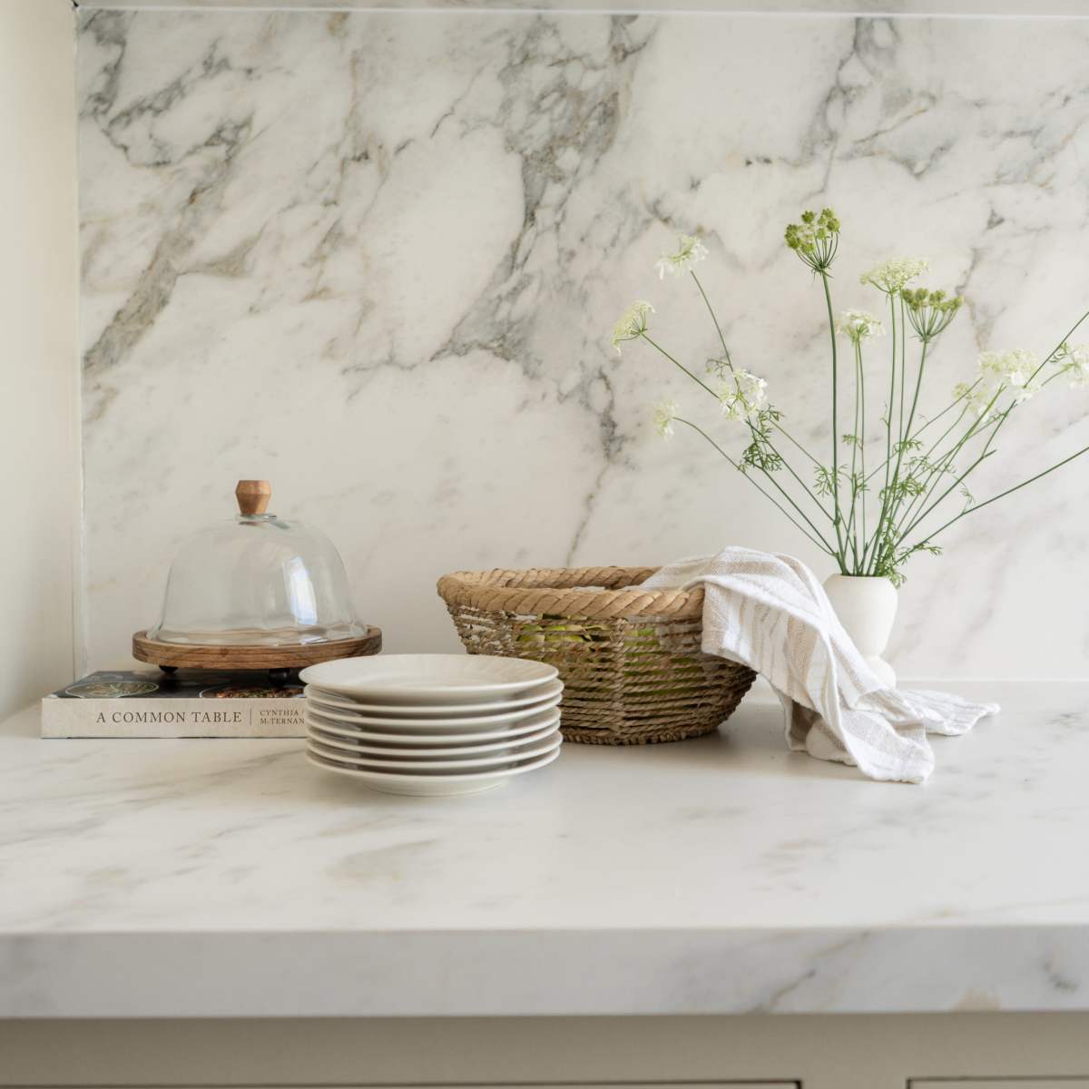 A marble countertop displays a stack of plates, a glass cloche, a woven basket, and a vase with flowers, with additional dishware arranged on a shelf above.