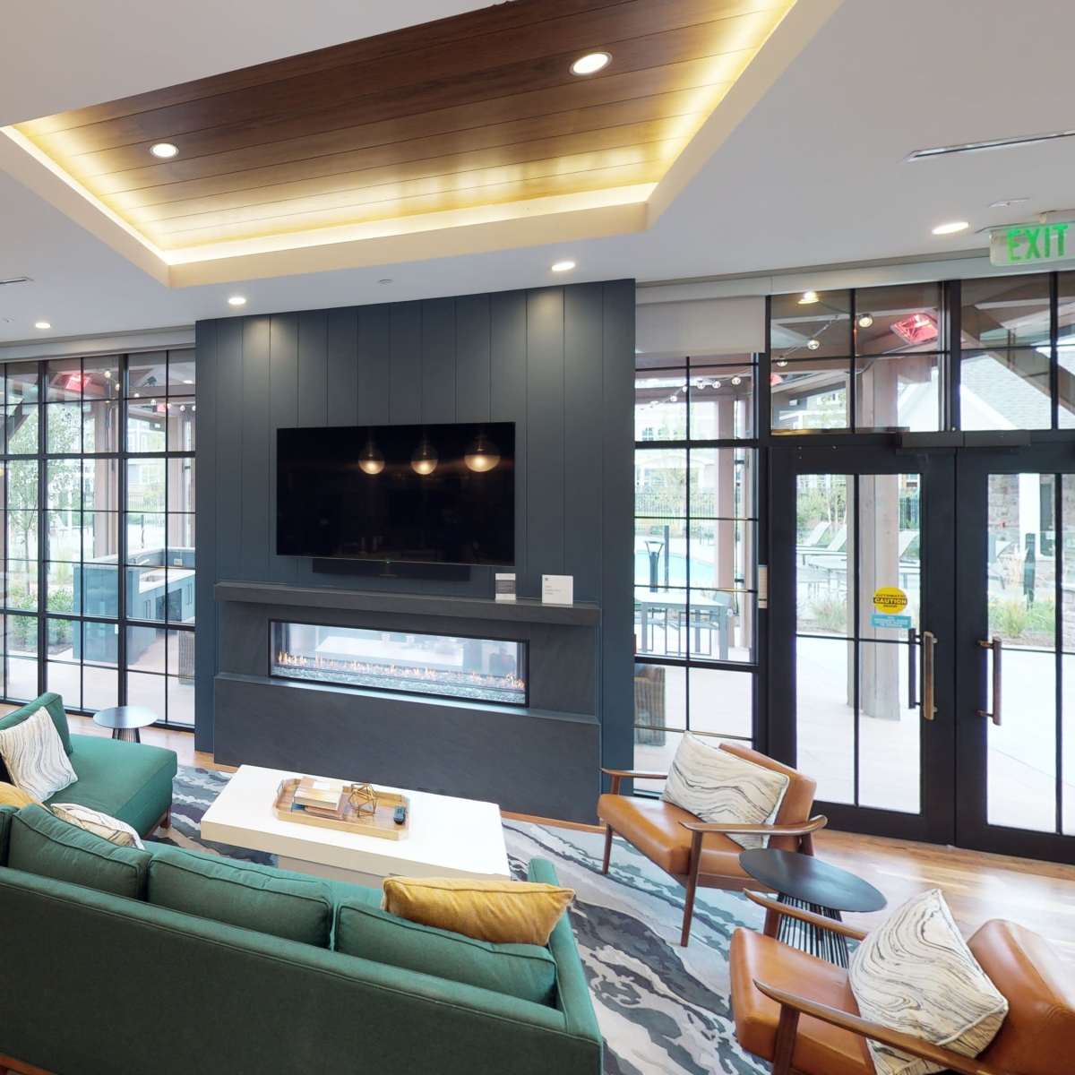 A modern lounge area with large windows, featuring a green sofa, a coffee table, an entertainment unit with a TV, and a mix of seating arrangements.