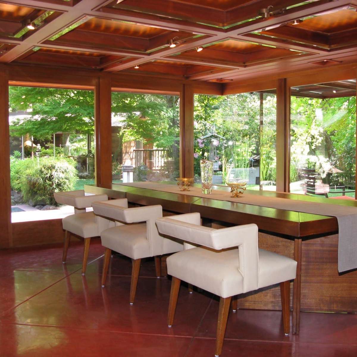 A modern wooden bar area features several cream-colored stools and large windows overlooking a lush green garden.