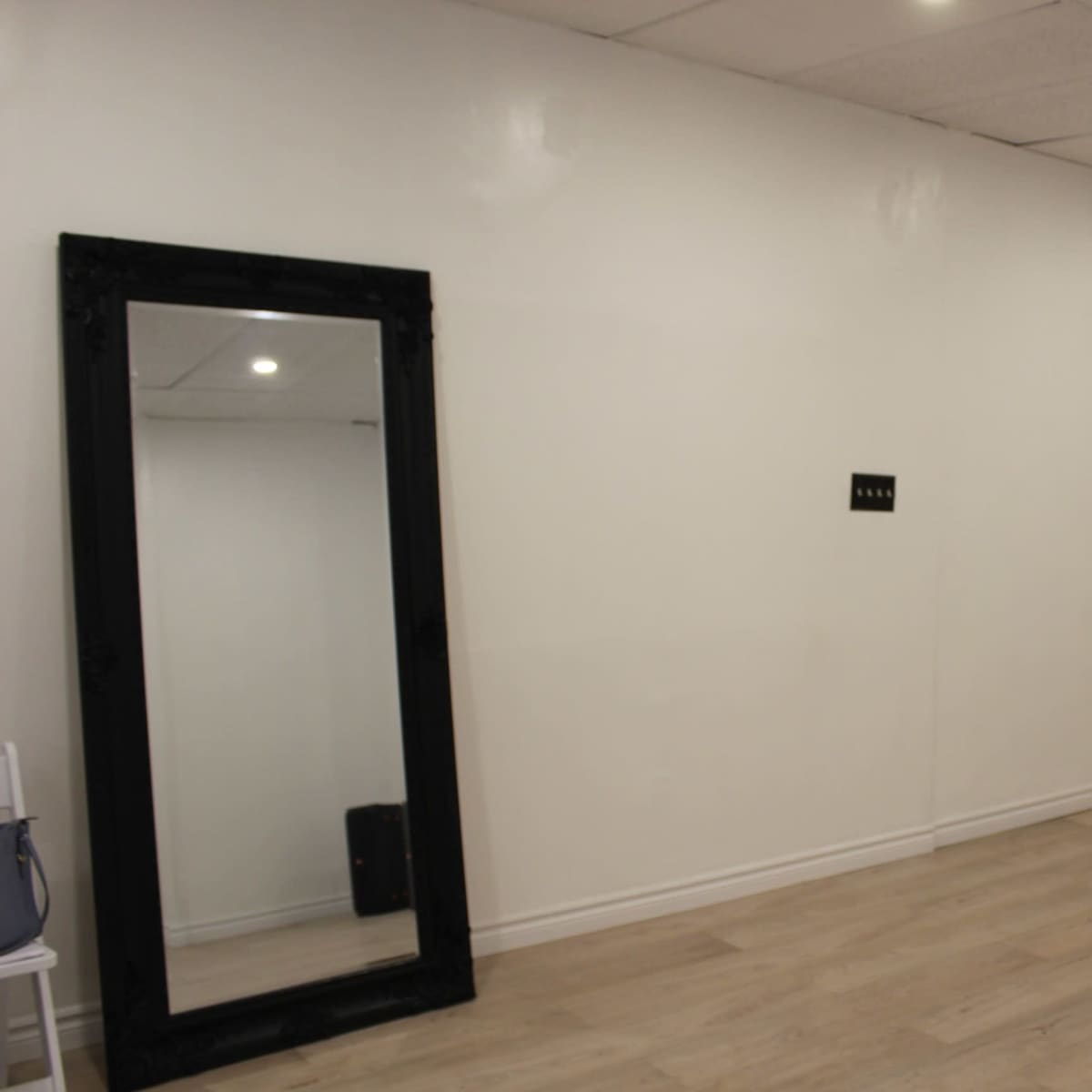 A spacious, empty room with light wooden flooring, white walls, and a large black-framed mirror on one side.
