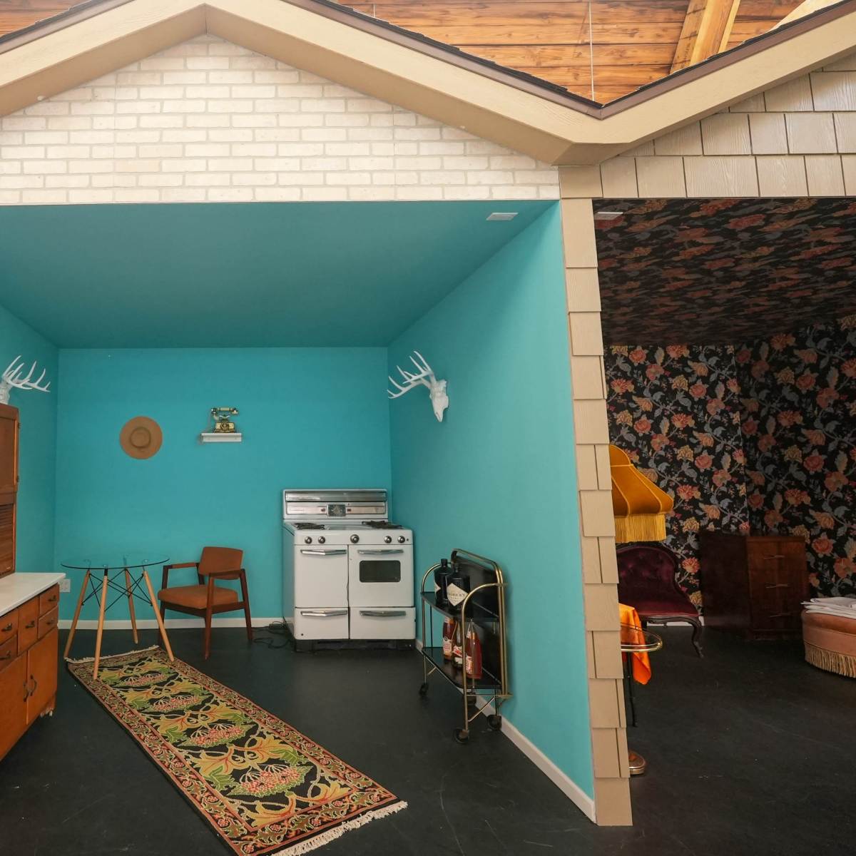 A brightly colored room designed to resemble a small house interior, featuring a turquoise wall, vintage furniture, and a decorative area rug.