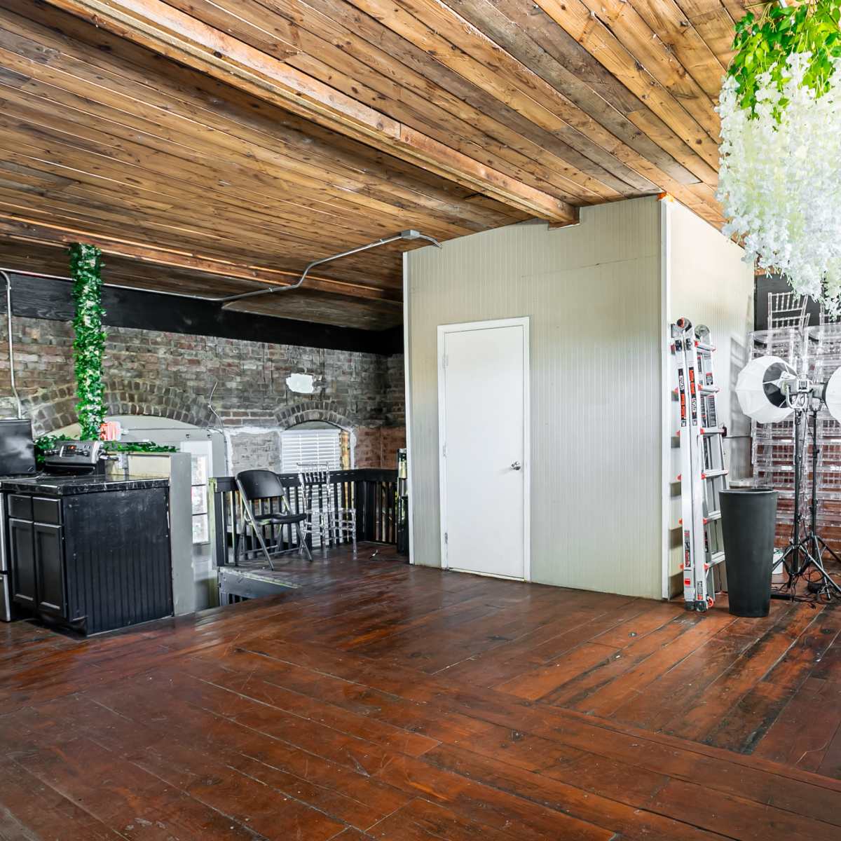 An industrial-style room with exposed brick walls, a wooden ceiling, and a divided space that includes a kitchenette area and photography equipment.