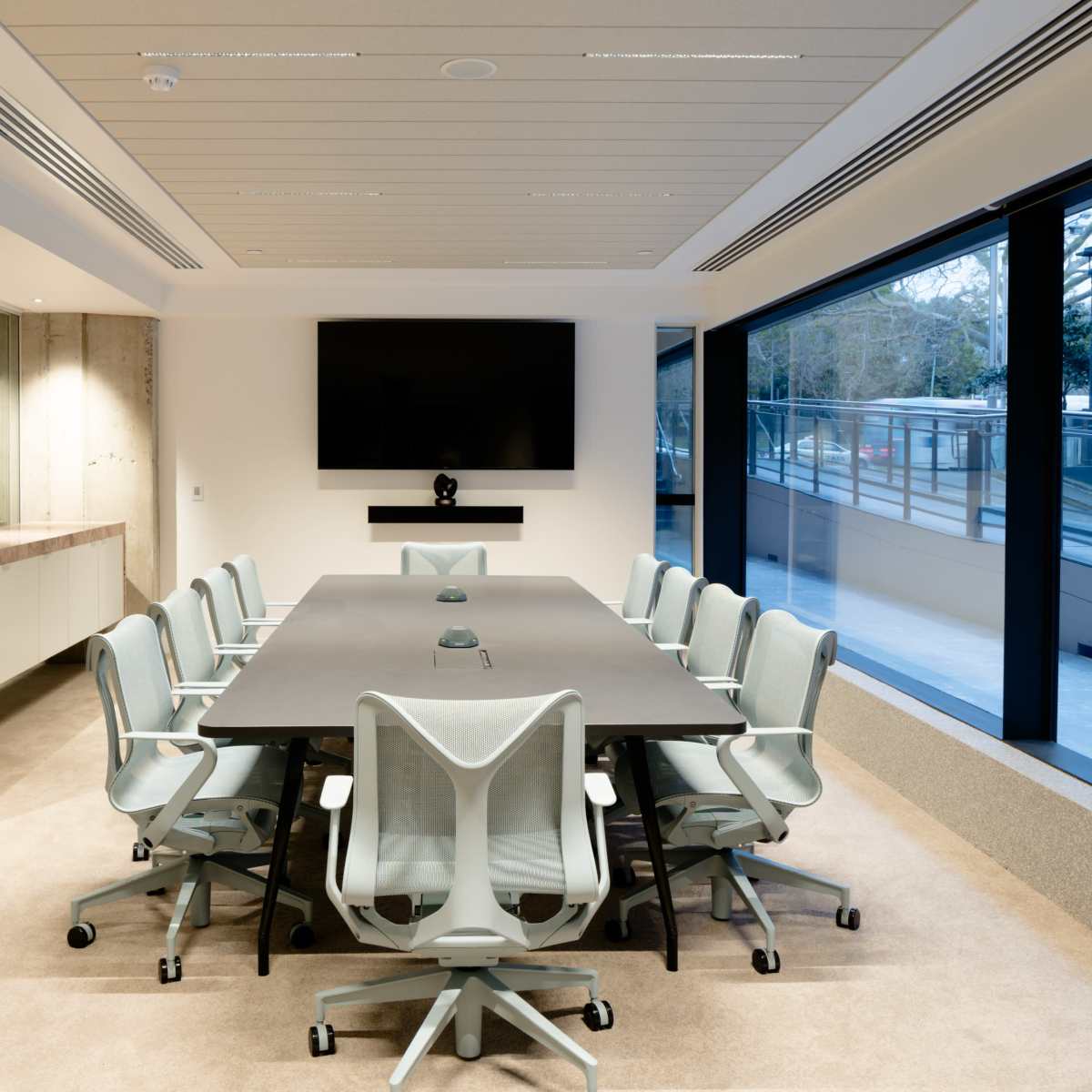 A modern conference room features a large rectangular table surrounded by ergonomic chairs, with a large screen mounted on the wall.