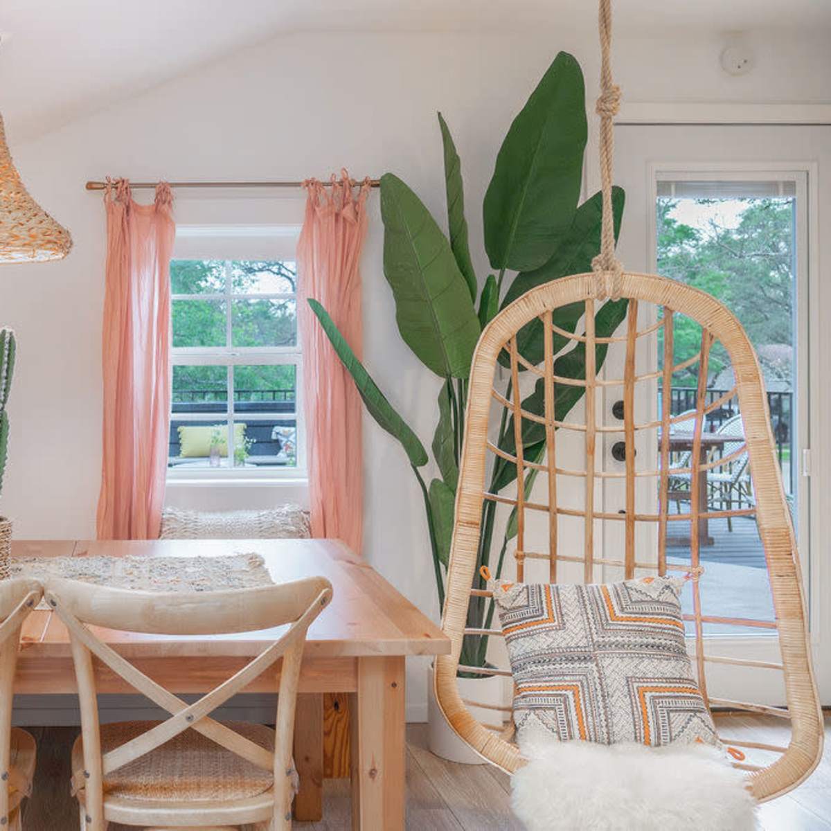 A light and airy dining area features a wooden table surrounded by chairs, a hanging rattan light fixture, a swing chair, and decorative plants.
